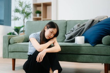 Depresyondaki Asyalı kadın oturma odasında kanepede oturmaktan endişe duyuyor. Hastalık, sağlık ve iyileşme kavramı.