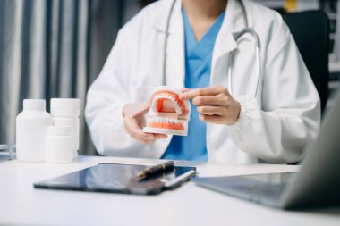 Diş hekimi, çene örnekleriyle masada oturan diş modeli ve diş sağlığı kliniğinde tablet ve dizüstü bilgisayarla çalışıyor.