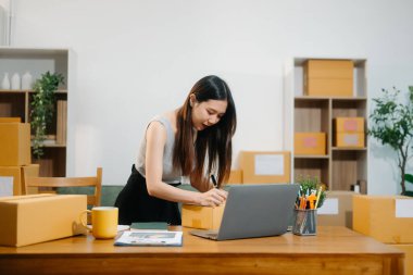 Startup small business SME, Entrepreneur owner woman using smartphone or tablet taking receive and checking online purchase shopping order to preparing pack product box. 