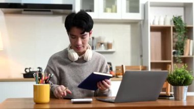 Asyalı genç adam dizüstü bilgisayar kullanırken ve not alırken evde çalışıyor.