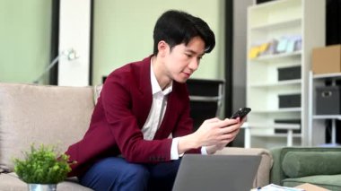 Kanepede otururken evde dizüstü bilgisayar ve akıllı telefonla çalışan genç bir işadamı.