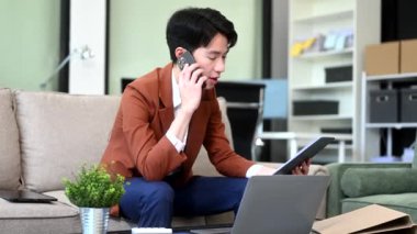 Genç işadamı evde dizüstü bilgisayarla çalışıyor ve kanepede otururken telefonla konuşuyor.