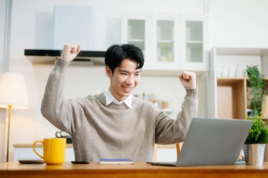 Genç adam evde otururken dizüstü bilgisayar kullanıyor. Genç adam mutfakta oturuyor ve dizüstü bilgisayarla çalışıyor.