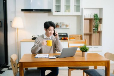 Genç adam evde otururken dizüstü bilgisayar kullanıyor. Genç adam mutfakta oturuyor ve dizüstü bilgisayarla çalışıyor.