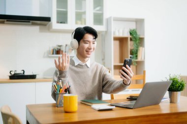 Genç adam evde otururken dizüstü bilgisayar kullanıyor. Genç adam mutfakta oturuyor ve dizüstü bilgisayarla çalışıyor.