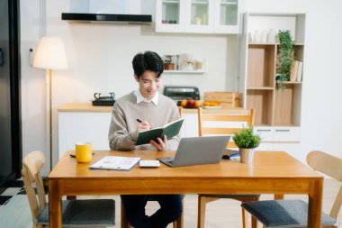 Genç adam evde otururken dizüstü bilgisayar kullanıyor. Genç adam mutfakta oturuyor ve dizüstü bilgisayarla çalışıyor.