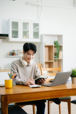 Genç adam evde otururken dizüstü bilgisayar kullanıyor. Genç adam mutfakta oturuyor ve dizüstü bilgisayarla çalışıyor.