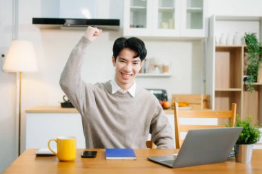 Genç adam evde otururken dizüstü bilgisayar kullanıyor. Genç adam mutfakta oturuyor ve dizüstü bilgisayarla çalışıyor.