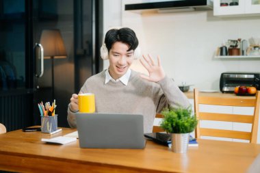 Genç adam evde otururken dizüstü bilgisayar kullanıyor. Genç adam mutfakta oturuyor ve dizüstü bilgisayarla çalışıyor.