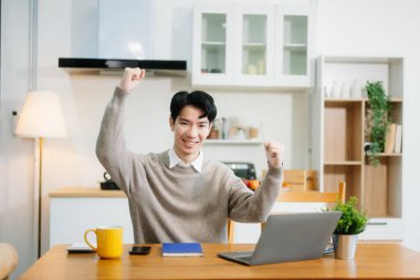 Genç adam evde otururken dizüstü bilgisayar kullanıyor. Genç adam mutfakta oturuyor ve dizüstü bilgisayarla çalışıyor.