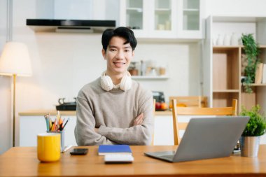 Genç adam evde otururken dizüstü bilgisayar kullanıyor. Genç adam mutfakta oturuyor ve dizüstü bilgisayarla çalışıyor.