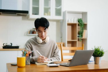 Genç adam evde otururken dizüstü bilgisayar kullanıyor. Genç adam mutfakta oturuyor ve dizüstü bilgisayarla çalışıyor.