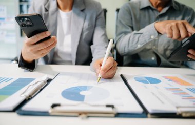 Ofis masasında akıllı telefon ve dizüstü bilgisayarı olan iş belgeleri ve arka planda sabah vakti veri tartışması yapan iki iş arkadaşı.