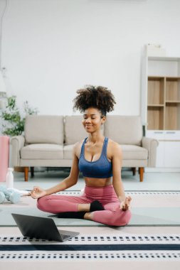 Genç bir kadın yoga yapıyor ve paspas ve jimnastik malzemeleriyle esniyor, sağlığını, motivasyonunu ve modern bir ev ortamında sağlıklı ve aktif bir yaşam tarzını gösteriyor.