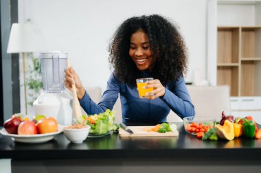 Modern bir mutfakta, taze meyve, sebze, blender ve portakal suyuyla gülümseyen bir kadın, temiz yeme, sağIık ve sağlıklı bir yaşam tarzını teşvik ediyor.
