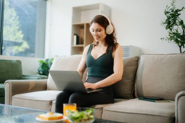 Kulaklıklı mutlu kadın yemek ve içeceklerle dolu bir oturma odasında dizüstü bilgisayar kullanıyor. Uzaktan çalışma, serbest çalışma, sağlıklı yaşam temaları için mükemmel.