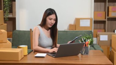 Bilgisayarda çalışan genç bir kadın. Evde kutular ve tabletlerle çalışıyor.