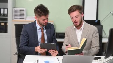 İki ofis arkadaşı dijital tablet ve dizüstü bilgisayar kullanarak, modern bir çalışma alanında bir başlangıç projesi hakkında tartışıyorlar.                       