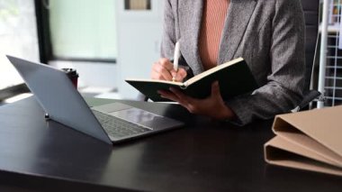 İş kadını ofis masasında dizüstü bilgisayar kullanıyor, not defterine not alıyor.