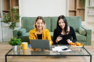 Yeşil kanepenin yanında oturan iki neşeli kadın şık bir evde dizüstü bilgisayar ve tablet kullanarak fikir alışverişinde bulunuyorlar. Takım çalışması çalışma alanı temaları için mükemmel