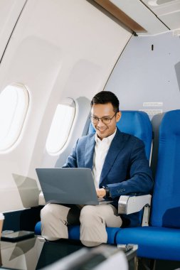 Çekici Asyalı erkek uçak yolcusu dizüstü bilgisayarla çalışırken rahat koltukta oturuyor. Stil içinde seyahat et, zarafetle çalış.