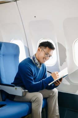 Asyalı bir adam uçak koltuğunda oturmuş, tatile giderken not defterine notlar yazıyor.. 