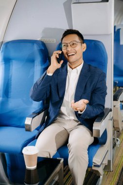 Akıllı telefonda konuşurken rahat koltukta oturan çekici Asyalı erkek yolcu. Stil içinde seyahat et, zarafetle çalış.