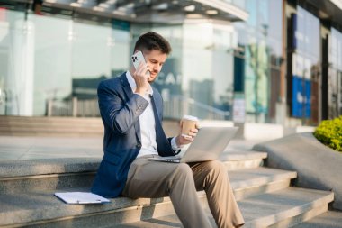 Modern iş adamı akıllı telefondan konuşuyor, dizüstü bilgisayar kullanıyor ve şehirdeki iş merkezinde kahve içiyor.