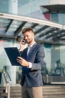 Modern iş adamı akıllı telefondan konuşuyor ve şehirdeki iş merkezinde pano tutuyor.