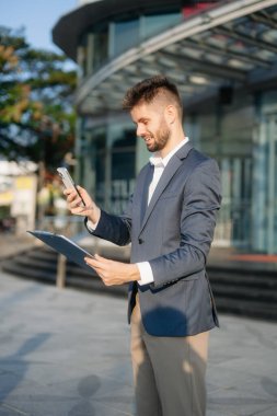 Modern iş adamı akıllı telefon kullanıyor ve şehirdeki iş merkezinde pano tutuyor.