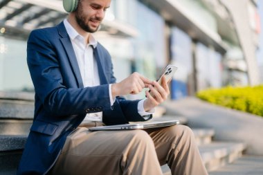 Modern iş adamı akıllı telefon kullanır ve şehirdeki iş merkezinde kulaklıkla müzik dinler.