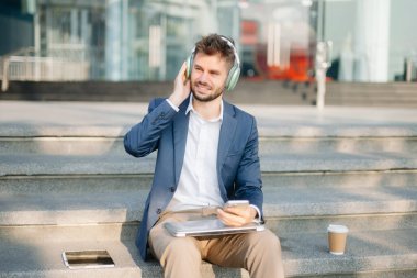 Modern iş adamı akıllı telefon kullanır ve şehirdeki iş merkezinde kulaklıkla müzik dinler.