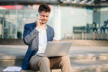 Modern iş adamı akıllı telefondan konuşuyor ve şehirdeki iş merkezinde dizüstü bilgisayar kullanıyor.