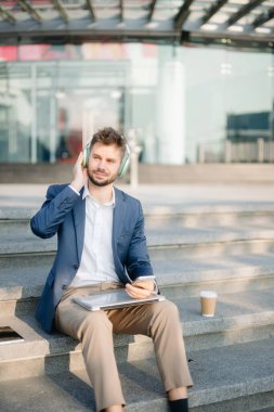 Modern iş adamı akıllı telefon kullanır ve şehirdeki iş merkezinde kulaklıkla müzik dinler.