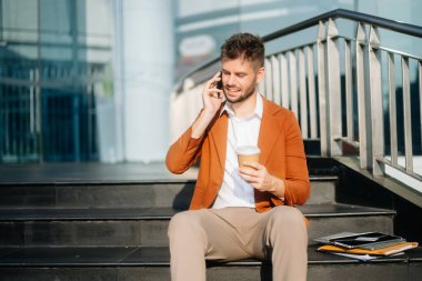 Modern iş adamı akıllı telefondan konuşuyor ve şehirdeki iş merkezinde kahve içiyor.