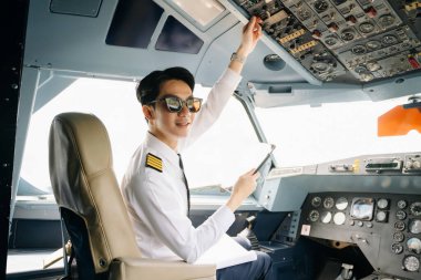 Asyalı kendinden emin üniformalı erkek pilot uçak kokpitinde oturuyor ve uçuş planlarını kontrol ediyor.