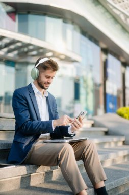 Modern iş adamı akıllı telefon kullanır ve şehirdeki iş merkezinde kulaklıkla müzik dinler.