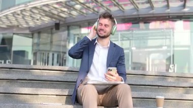 Şehirdeki merdivenlerde oturan genç adam akıllı telefonuyla dizlerinde dizüstü bilgisayarla kulaklıkla müzik dinliyor.