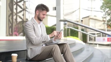 Genç işadamı şehirde merdivenlerde otururken dizlerinde dizüstü bilgisayarla akıllı telefon kullanıyor.