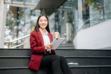Ofisin dışında elinde tablet tutarken gülümseyen ve kahve içen kendine güvenen Asyalı kadın. İş merkezi arka planında büyük şehir