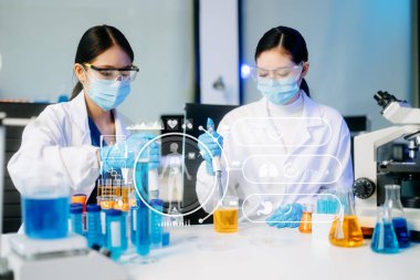 Yüz maskeli iki Asyalı kadın bilim adamı yüksek teknolojili bir tıp laboratuarında mikro pipetler kullanarak örnekleri analiz ederek biyokimyasal araştırma yapıyorlar. Tıbbi simgeleri olan sanal ekran 