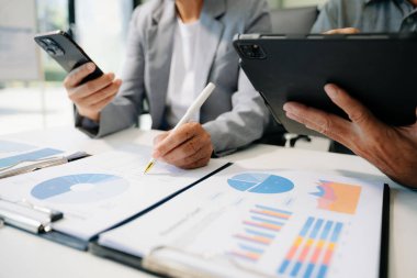 Ofis masasında akıllı telefonlu iş belgeleri ve arka planda sabah vakti veri tartışması yapan iki iş arkadaşı.