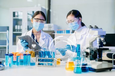 Yüz maskeli iki Asyalı kadın bilim adamı yüksek teknolojili bir tıp laboratuarında mikro pipetler kullanarak örnekleri analiz ederek biyokimyasal araştırma yapıyorlar. Tıbbi simgeleri olan sanal ekran 