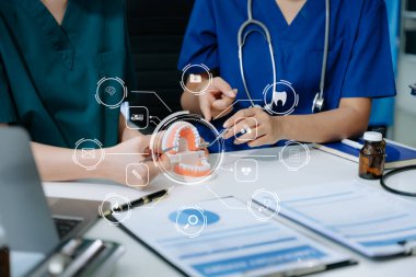 Tıbbi simgeler ve iki dişçi çene örnekleriyle masada oturuyor diş modeli ve diş sağlığı kliniğinde dizüstü bilgisayarla çalışıyor. 