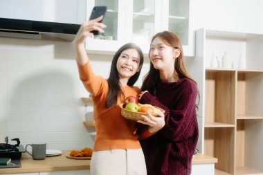 İki genç Asyalı kadın sıcacık bir mutfakta selfie çekiyor, gülümsüyor, sohbet ediyor ve sıcak, rahat bir atmosferde elmaları paylaşıyorlar.. 