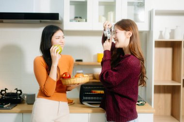 Sıcak bir mutfakta elma yiyen iki genç Asyalı kadın, biri diğerinin sıcak, rahat bir atmosferde fotoğrafını çekiyor..  