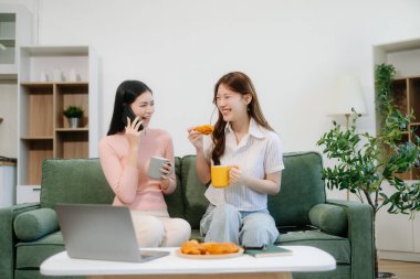 İki neşeli kadın yeşil bir kanepede oturmuş akıllı telefon kullanıyor ve şık bir evde atıştırmalık şeyler yiyorlar. Takım çalışması çalışma alanı temaları için mükemmel