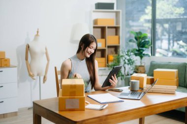 Startup small business SME, Entrepreneur owner woman using smartphone or tablet taking receive and checking online purchase shopping order to preparing pack product box.