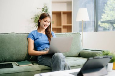 Genç Asyalı kadın evinde rahat bir kanepede rahat rahat oturup akıllı telefon ve tablet gülümseyen kızla mesajlaşırken evden cep telefonu alışverişini kullanıyor.