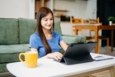 Modern evdeki kanepede otururken akıllı telefon ve tablet kullanan genç Asyalı kadın.